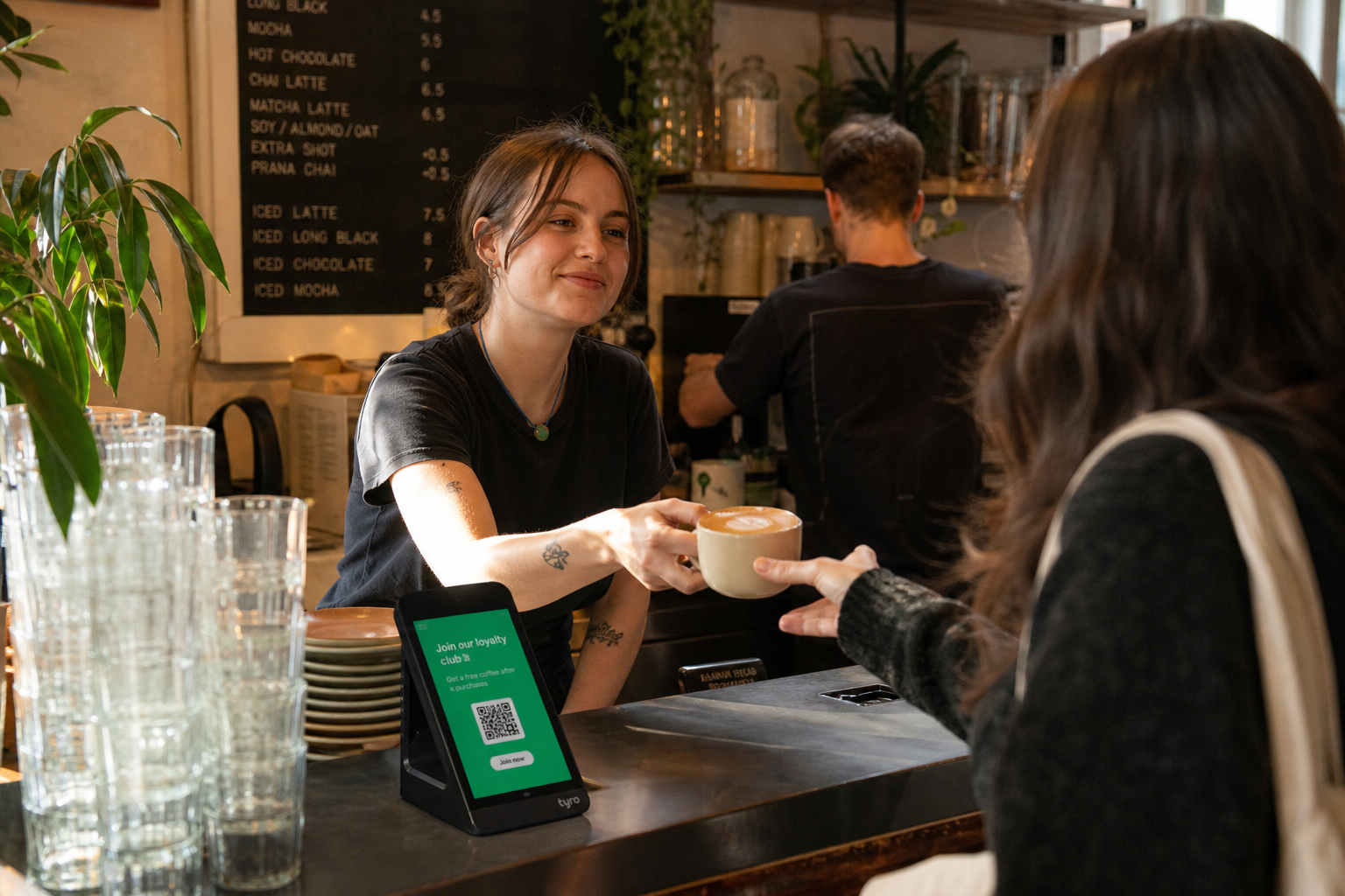 A barista handing a flat white across a busy café counter with a phone-based loyalty prompt on the card reader
