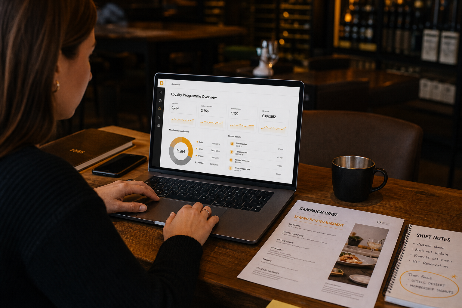 A hospitality marketing manager looking at a laptop with a loyalty dashboard open alongside campaign briefs and shift notes